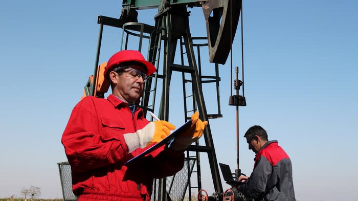 Two men writing in notebooks with an oil well in the background