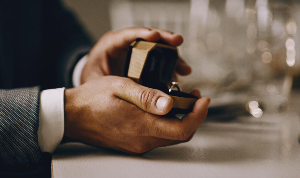 A hand holding an open ring box with a ring inside, suggesting a marriage proposal