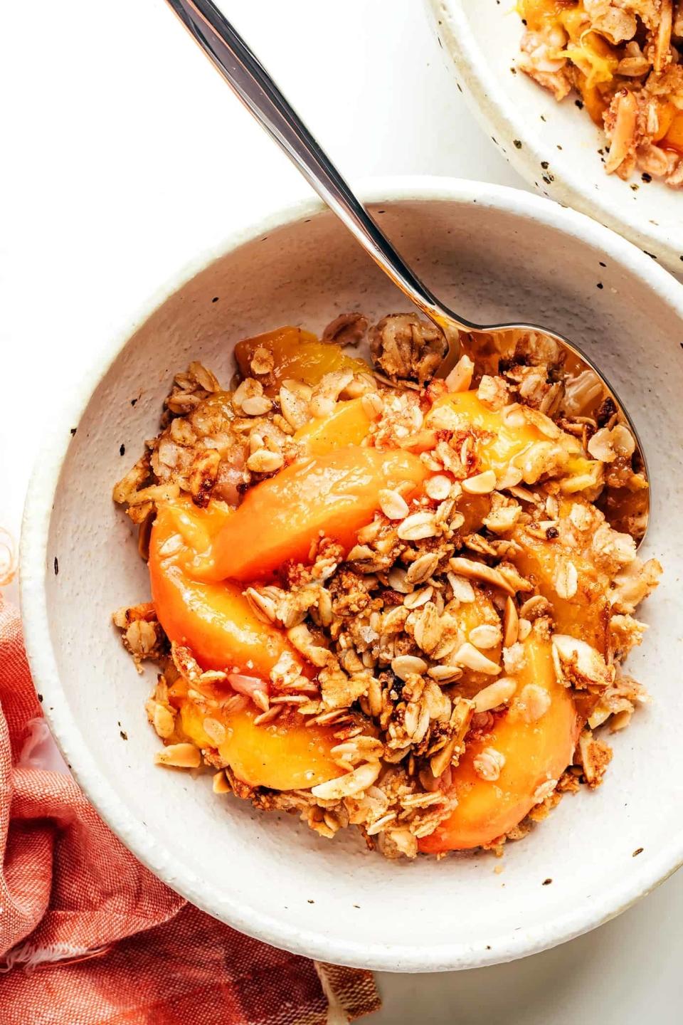 A bowl of peach crisp with a spoon, showing slices of peaches and a topping of oats and nuts. A napkin is partially visible beside the bowl