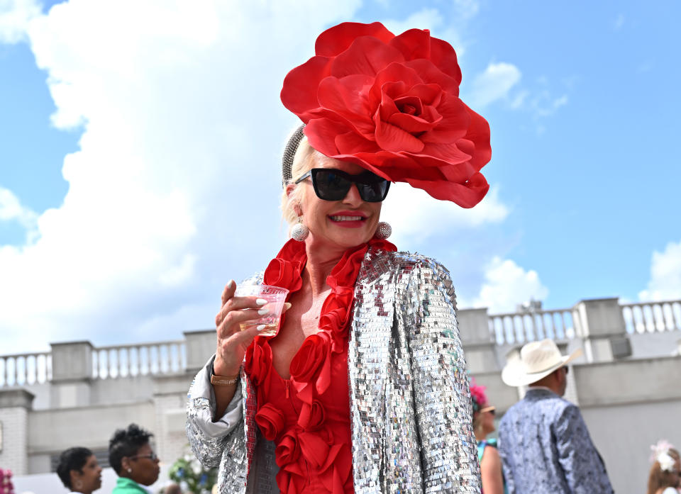 A woman in large sunglasses holds a drink while wearing a hat that looks like an enormous red flower.