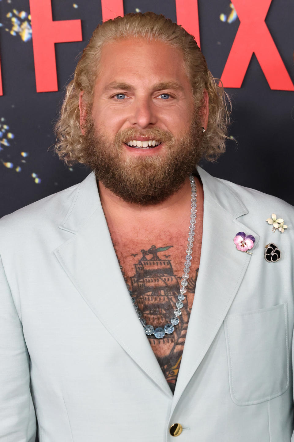 Jonah Hill, wearing a light-colored suit with a necklace and floral brooches, poses on the red carpet. He has a beard and curly hair