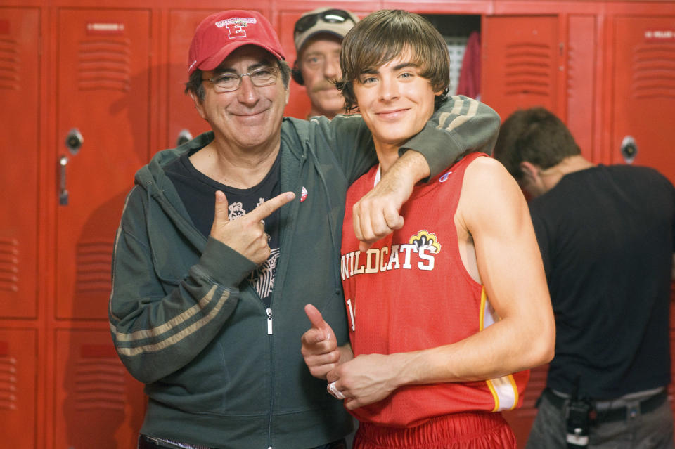 Kenny Ortega points beside Zac Efron in a Wildcats basketball jersey. A person stands blurred in the background near red lockers