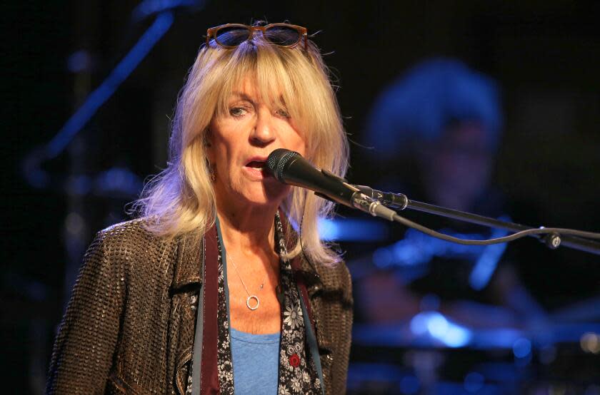 Christine McVie sings into a mic while wearing a brown jacket and blue shirt with sun glasses atop her head