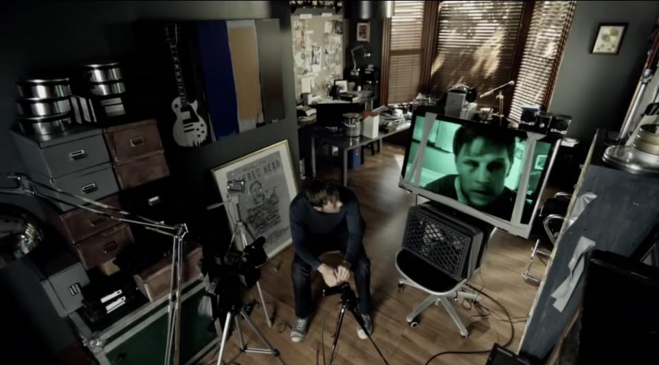 A man sitting in the middle of a room with a camera, TV screen, and other gadgets