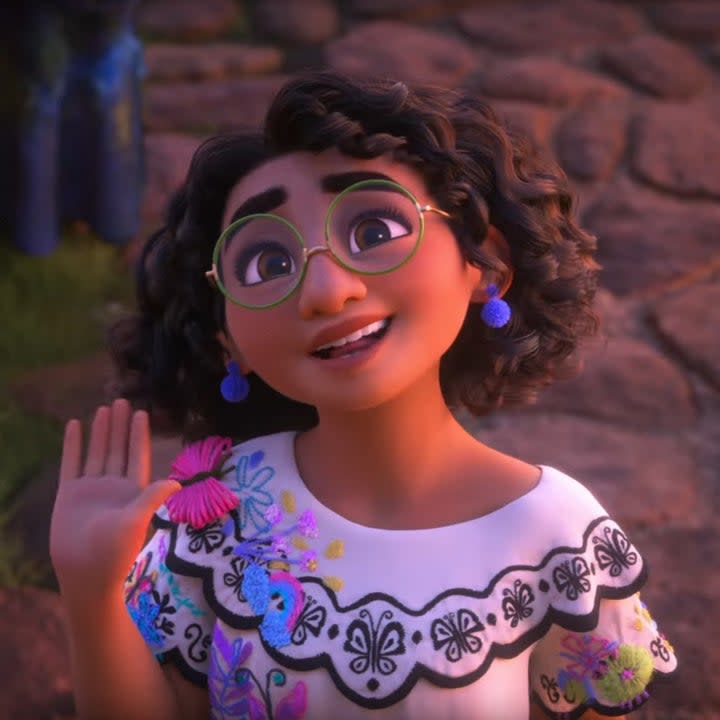 Mirabel wearing round eyeglasses and an embroidered dress decorated with butterflies while waving