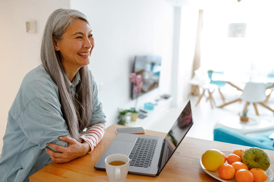 Joyful female person looking away and laughing while using modern notebook in kitchen