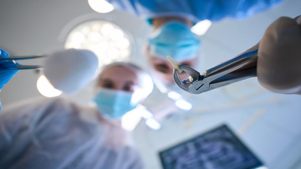 Dental surgeon standing over a person holding a tooth that has been extracted using forceps while another dentist stands next to them