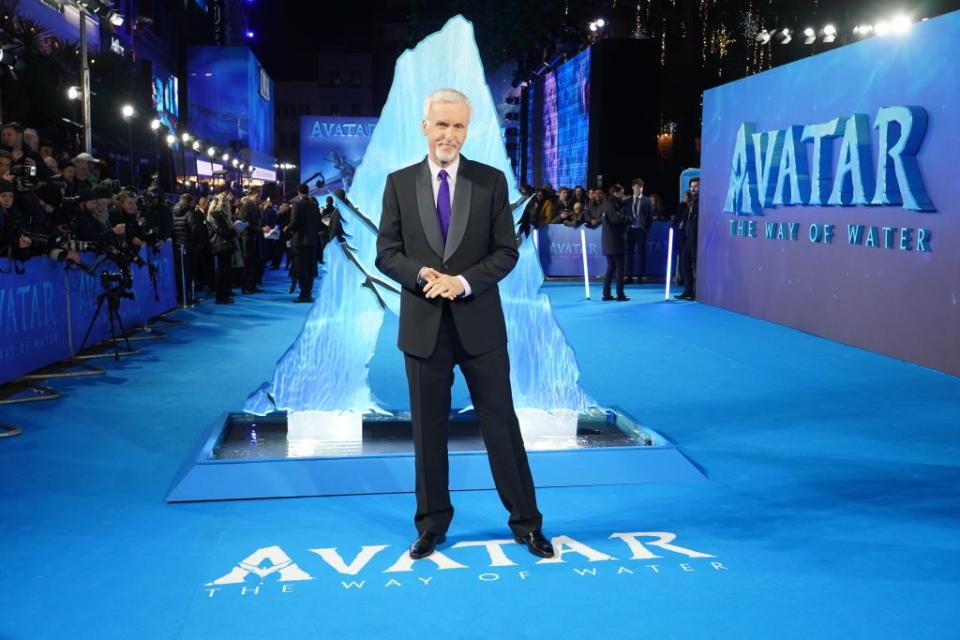 james cameron wearing a black suit and purple tie, standing in front of promotional material for avatar the way of water