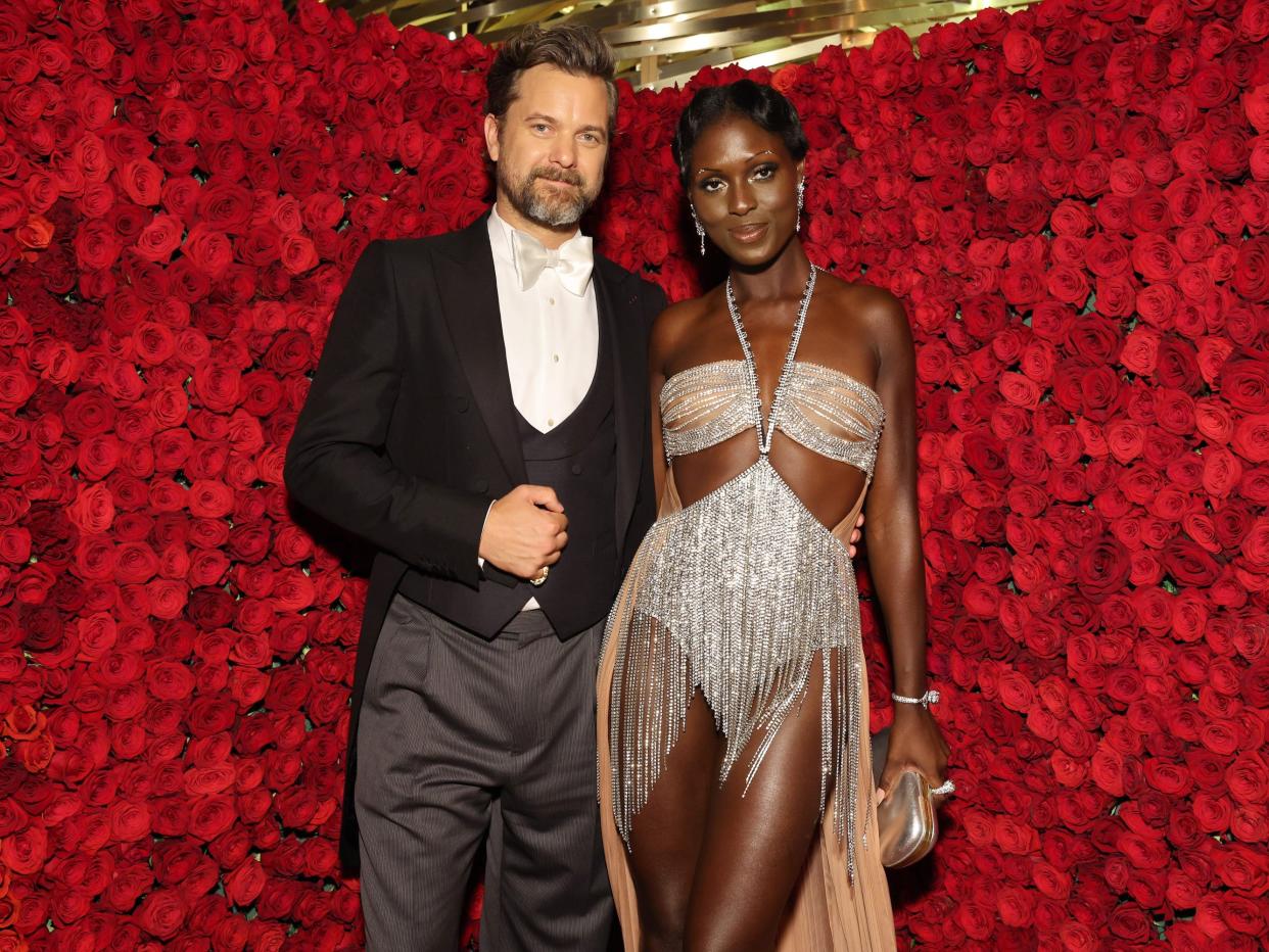 Joshua Jackson, in a black jacket with tails, and Jodie Turner-Smith, in a sparkly silver bodysuit, pose in front of a background of roses at the 2022 Met Gala.