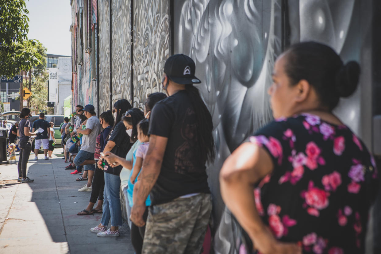 People stand in line against a shaded wall along a city sidewalk.