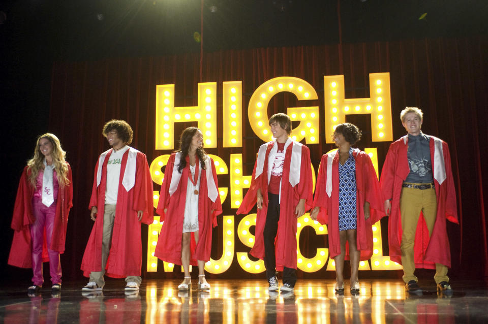 Ashley Tisdale, Corbin Bleu, Vanessa Hudgens, Zac Efron, Monique Coleman, and Lucas Grabeel stand in red graduation robes on stage in front of "High School Musical" sign