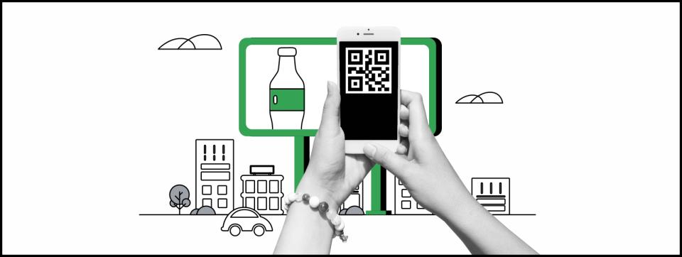 Unas manos en blanco y negro con un telefóno en la mano escanean un QR de un aviso de vía pública. Detrás un cartel en verde con una botella con etiqueta verde y el perfil de una ciudad.