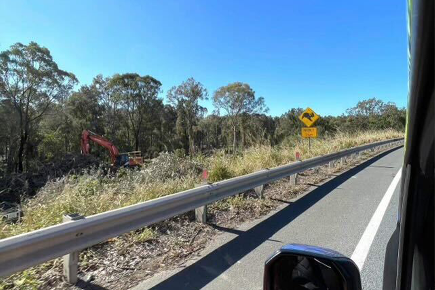 Stopped on the side of the road, and looking through a car window, we see a bulldozer in the distance, and a koala crossing sign a little closer by the side.