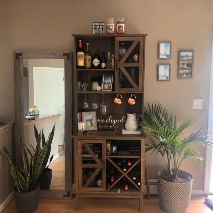 A wooden storage cabinet with various bottles of alcohol, glasses, and decor items. Nearby are a mirror, houseplants, and wall pictures