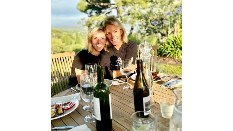 Two women smiling outside in the sun drinking wine 