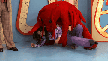 Person in plush red lobster costume over another person on a set with oversized letters in the background