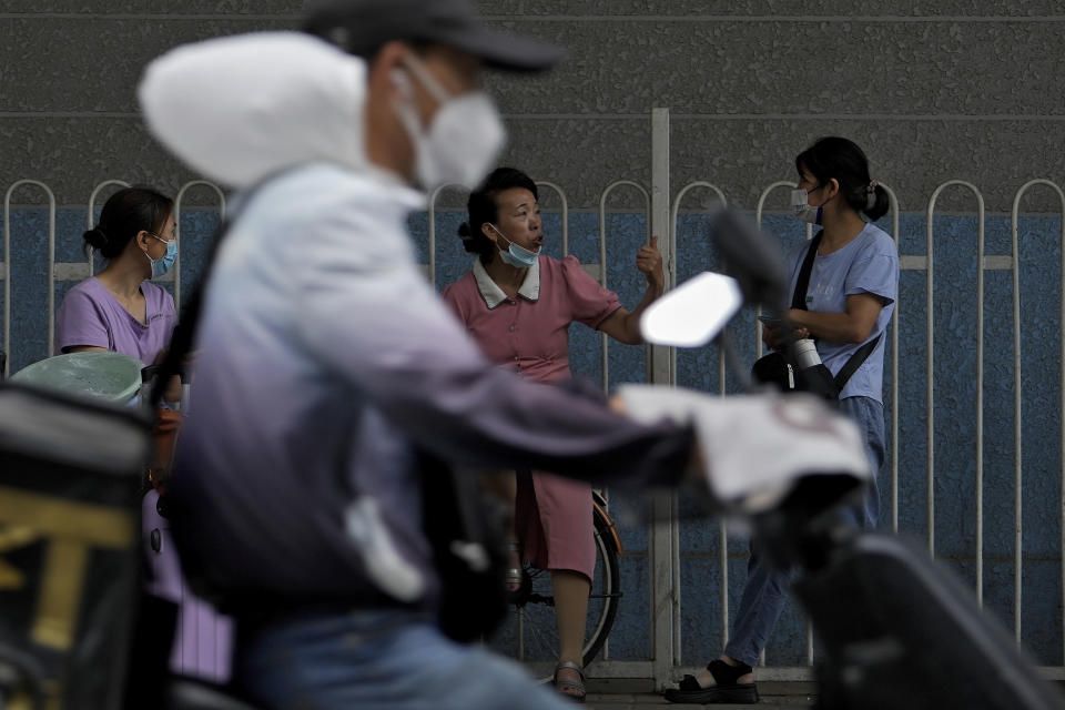 A delivery driver rides past women wearing face masks chatting with each other at a bus station in Beijing, Tuesday, June 28, 2022. (AP Photo/Andy Wong)