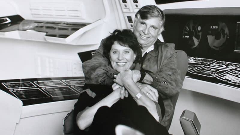 A smiling elderly man embraces a smiling woman in a white room with several dark computer consoles.