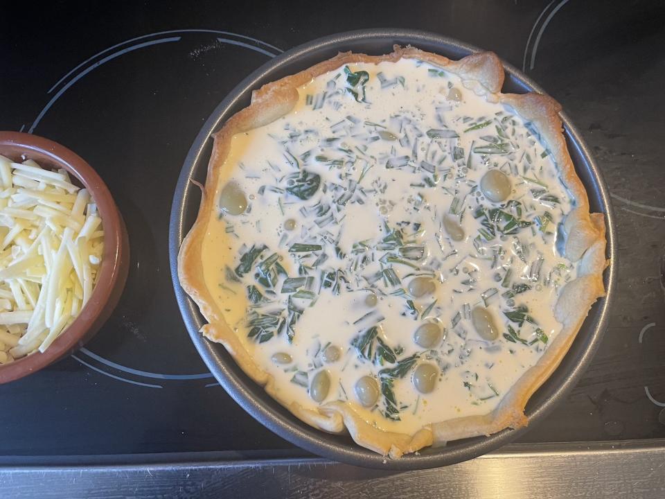 An overhead view of the uncooked quiche, with a pot of cheese about to be sprinkled on top.