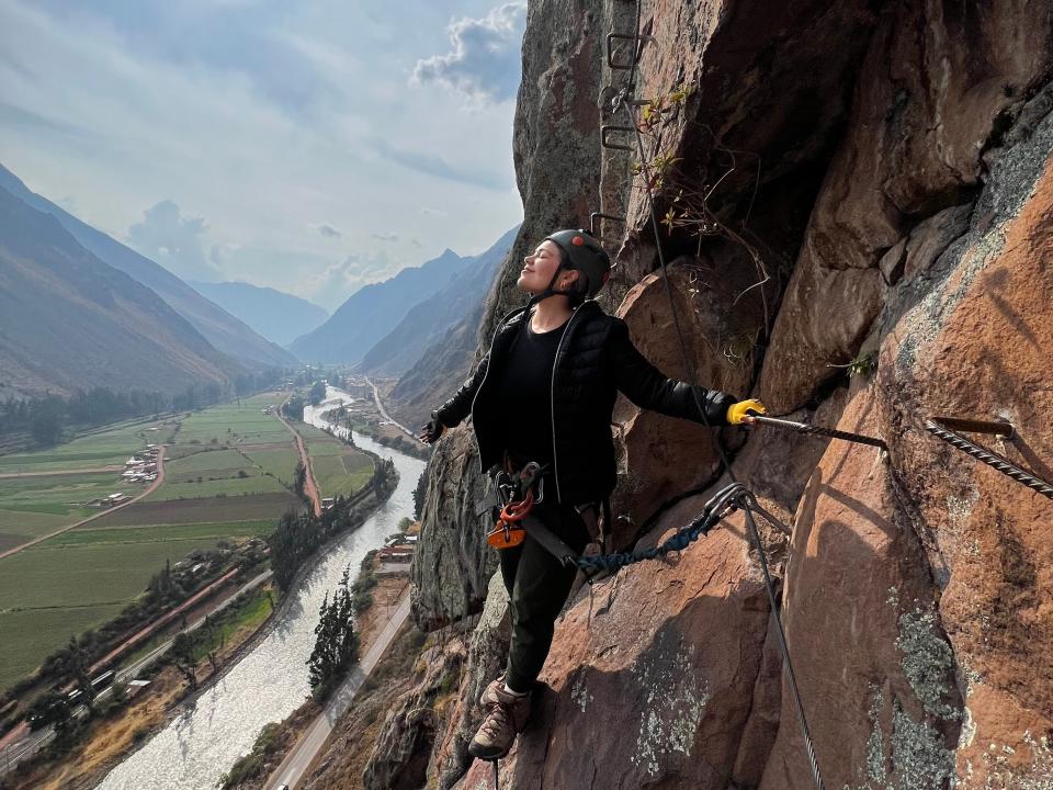 Victoria smiles with her eyes closed, looking away from the cliff as she climbs via ferrata.