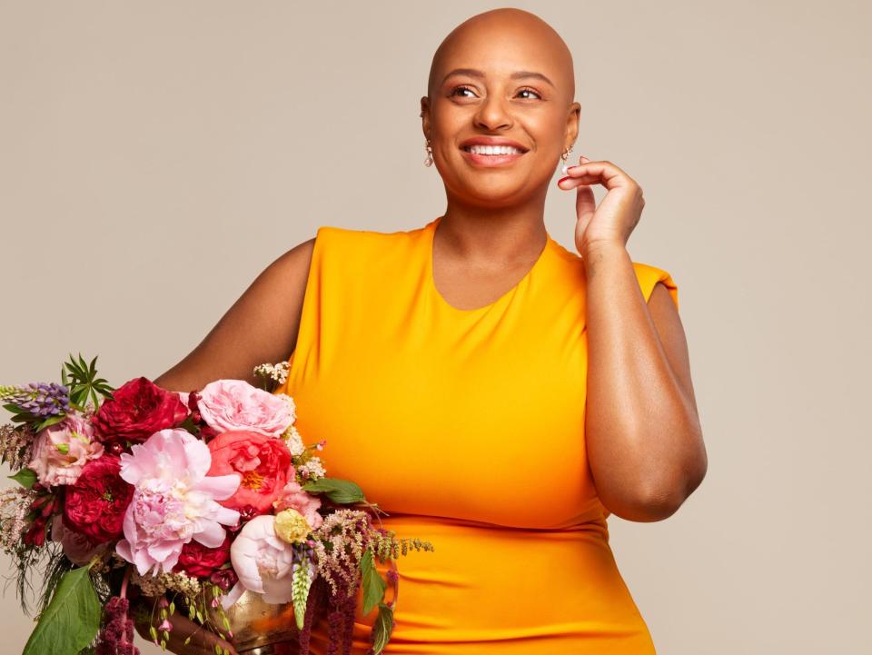 Christala Fletcher is bald, has dark eyes, and wears a mustard-colored sleeveless dress and is smiling off to the viewer's left. She poses her left hand by her ear and holds an arrangement of flowers in the other hand.
