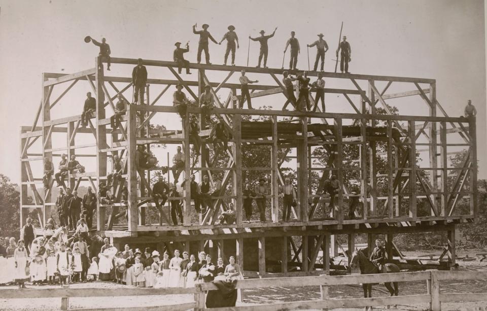 A 19th-century black-and-white image shows dozens of people crowded around the frame of a rising barn in Ohio