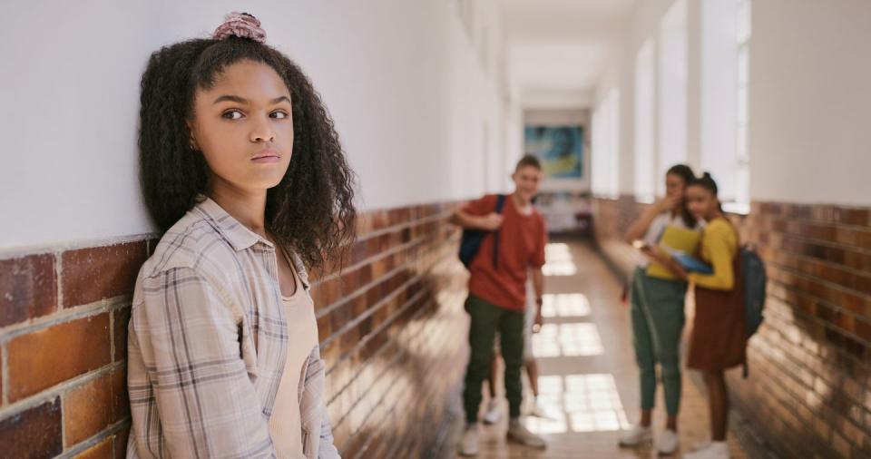 teen, young and lonely girl standing in the corridor at break time shy, depressed and unhappy child inside educational building sad youth looks away from students that are bullying her