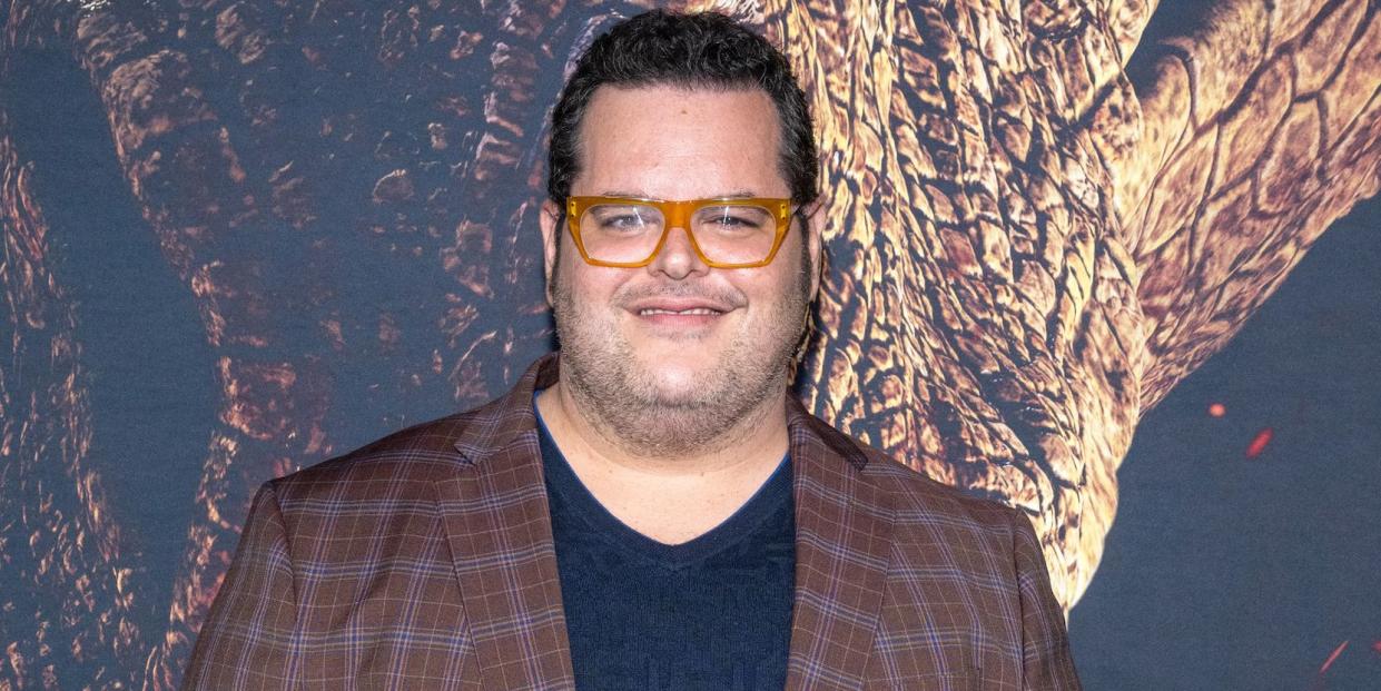 josh gad, a man stands smiling and looking at the camera, he wears a blue tshirt and jeans with a brown jacket and orange glasses