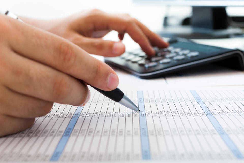 A person points a pen to a sheet with various numbers on it, with his other hand on a calculator.