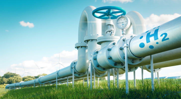 An image of a hydrogen fuel pipeline running through a grassy field, with wind turbines and solar panels in the background. top hydrogen stocks to buy