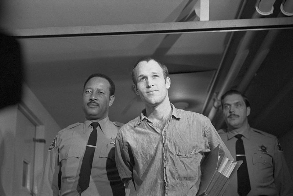 a man smiles slightly as two police officers escort him in handcuffs down a hallway