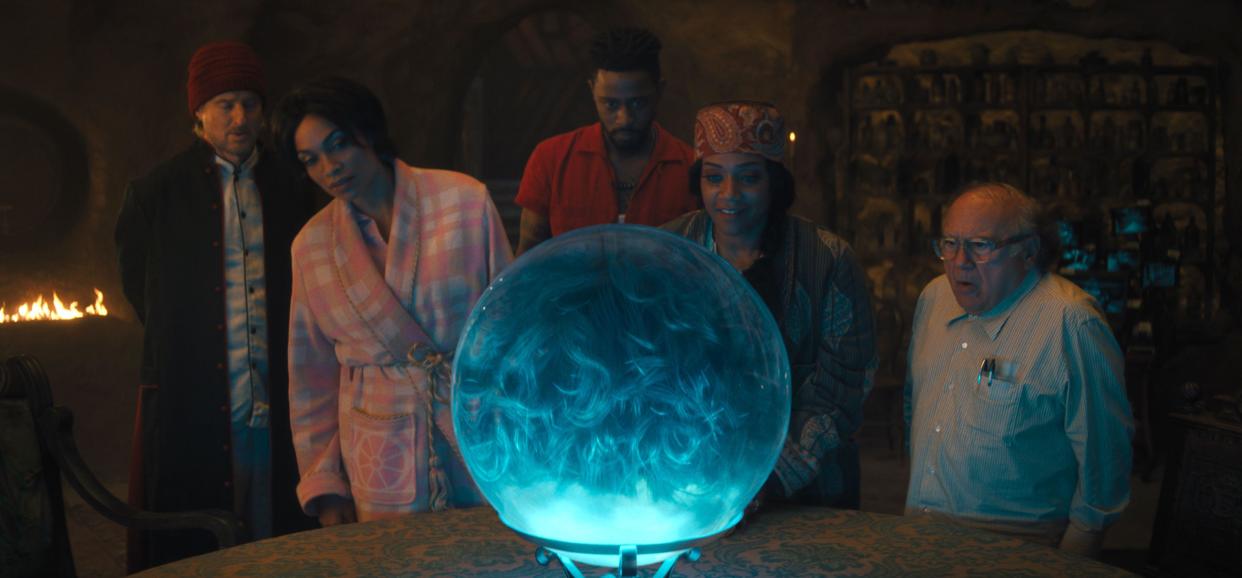  From left to right: Father Kent (Owen Wilson), Gabbie (Rosario Dawson), Ben Matthias (LaKeith Stanfield), Harriet (Tiffany Haddish) and Bruce Davis (Danny DeVito) stand around a table with a crystal ball on it in Haunted Mansion.  . 