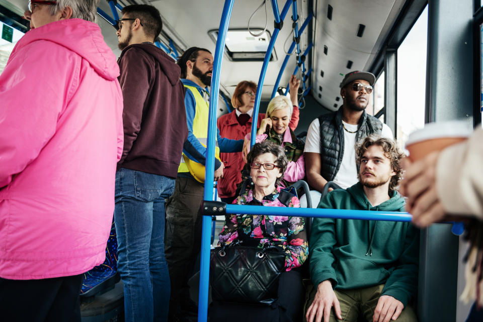 A group of people ride a crowded bus, with a woman in a floral top seated and others standing around her, looking at their phones or talking