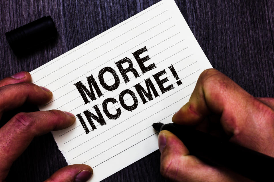 Two hands have written the words "MORE INCOME!" on an index card.