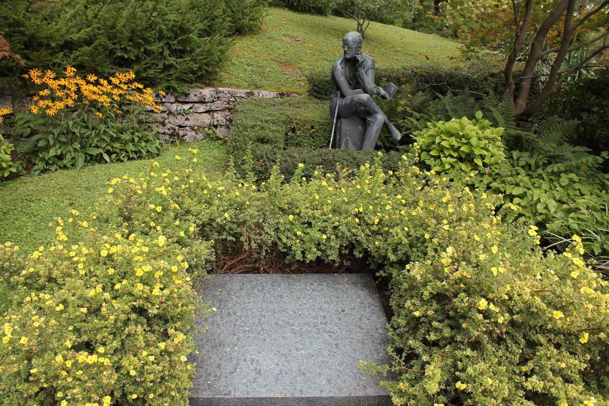 James Joyce's grave in the Fluntern Cemetery, Zurich, surrounded by yellow flowers in bloom with a statue of joyce in the background with many other plants and flowers