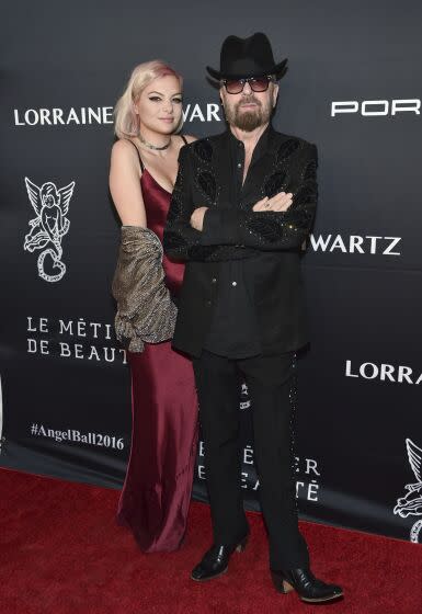 Kaya Stewart in a burgundy gown standing behind Dave Stewart in a black hat, sunglasses and a black suit