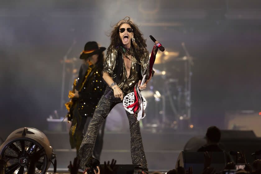 A man with long hair sings onstage with a guitarist and drummer behind him