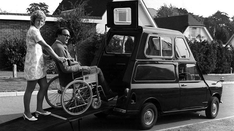 A vintage black-and-white photo of a woman helping a man in a wheelchari get into a custom made car.