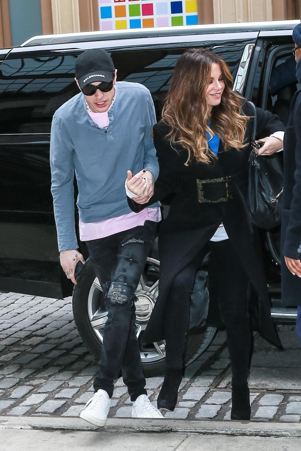 Pete Davidson in casual attire and Kate Beckinsale in a stylish black coat walk hand-in-hand outside a vehicle on a cobblestone street