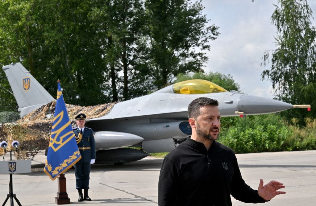 Ukraine's president stands in front of an F-16 at a press event. There are trees in the background, and the fighter jet is partially covered.