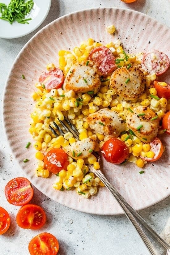 A plate of scallops topped with black pepper, served with corn, cherry tomatoes, and chives. A fork is placed on the plate