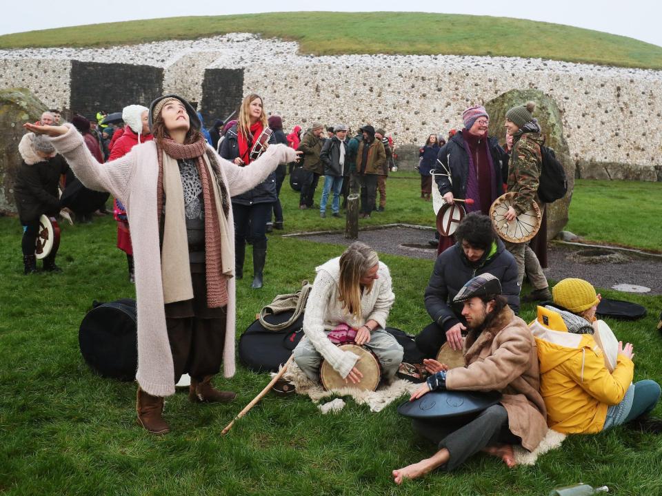 Peopel are sat on the floor or looking up at the sky. In the background, Newgrange appears as circular structure made of rocks surmounted by grass