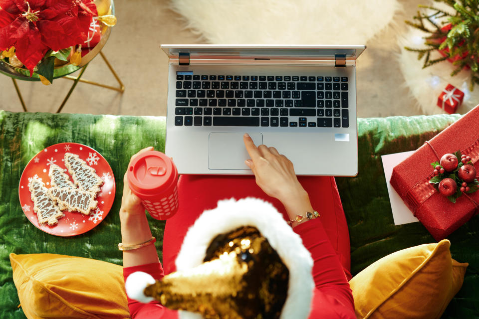 Holiday season. Upper view of young housewife in sparkle gold sequin santa hat with cup of coffee reading on a laptop while sitting on couch in the modern living room at Christmas.