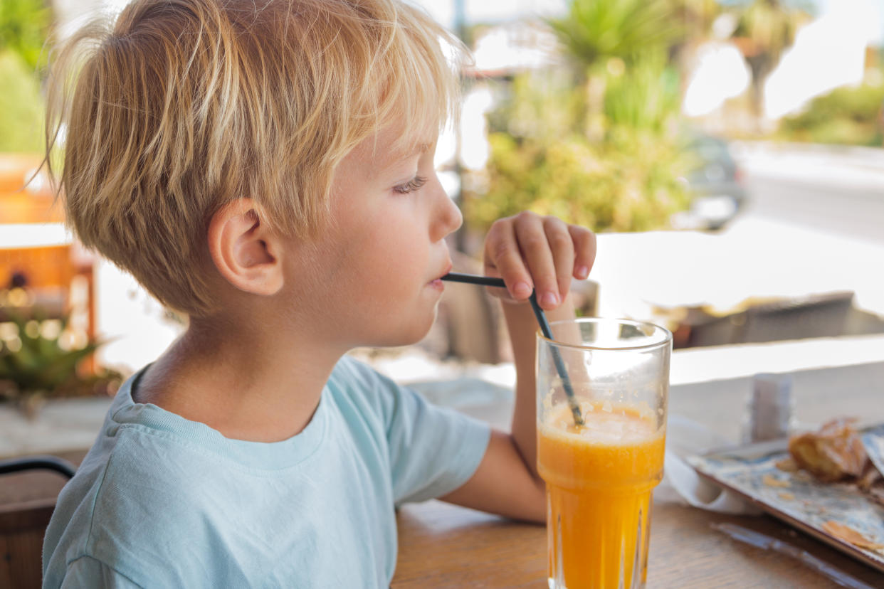 Joyful small boy (4-5 years) drinking fresh juice throw plastic straw. Happy preschool boy holding glass of orange juice with a plastic drinking straw.