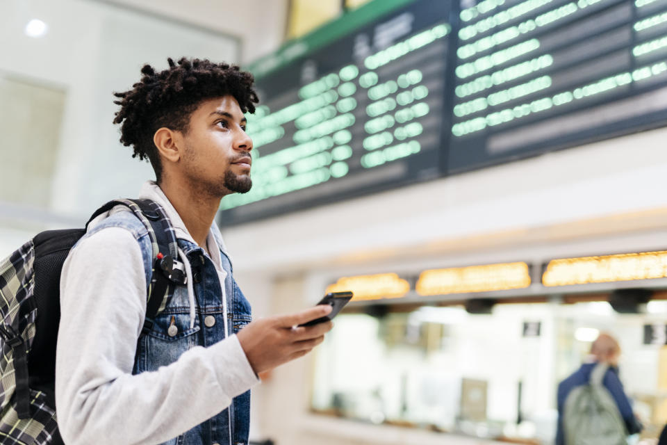 A person looks up at a departure board in a train station, holding a phone and wearing a denim jacket over a hoodie with a backpack