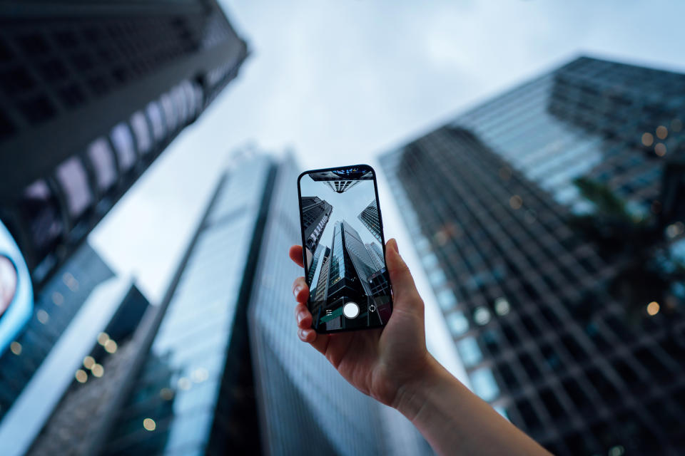 Personal perspective of woman's hand taking photos of urban skyscrapers in downtown district with smartphone, surrounded by modern and compact commercial buildings in the city