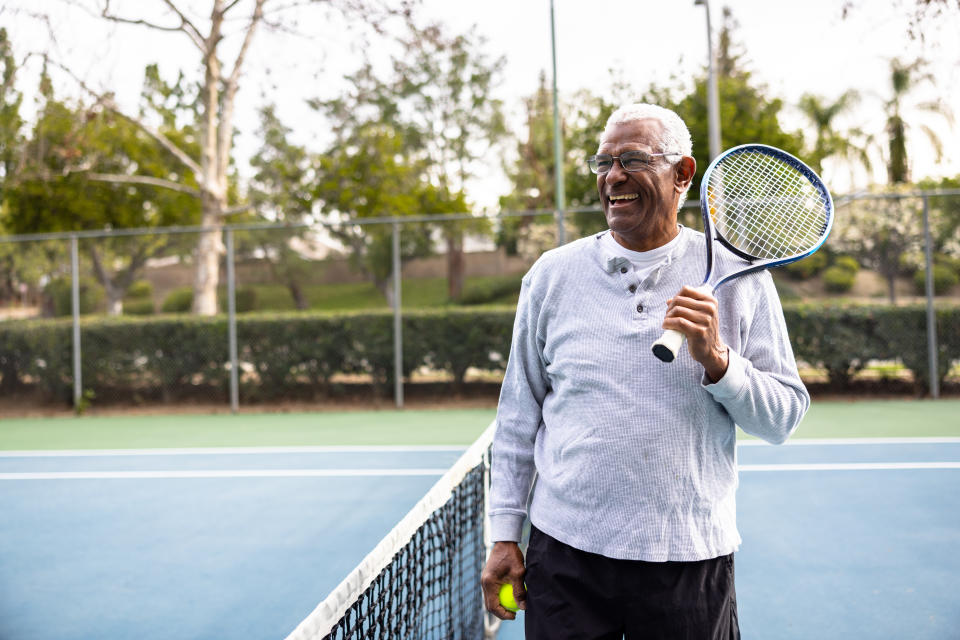 Older person with glasses and sports attire smiling while holding a tennis racket on a tennis court