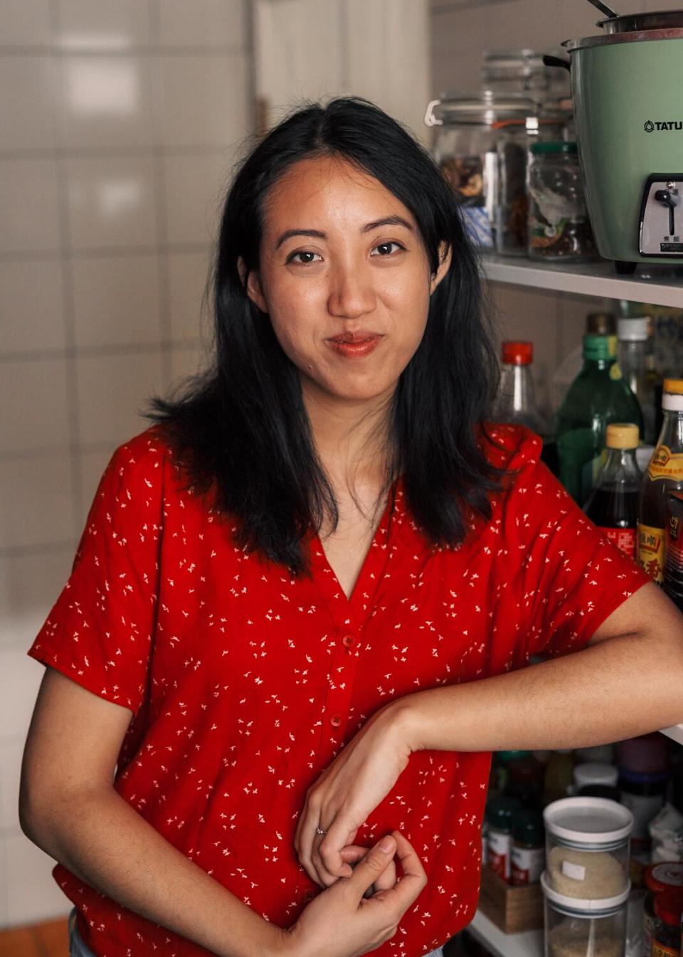 A woman in an orangish-red top stands in a kitchen.