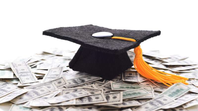A mortarboard sits on a pile of U.S. hundred-dollar bills.