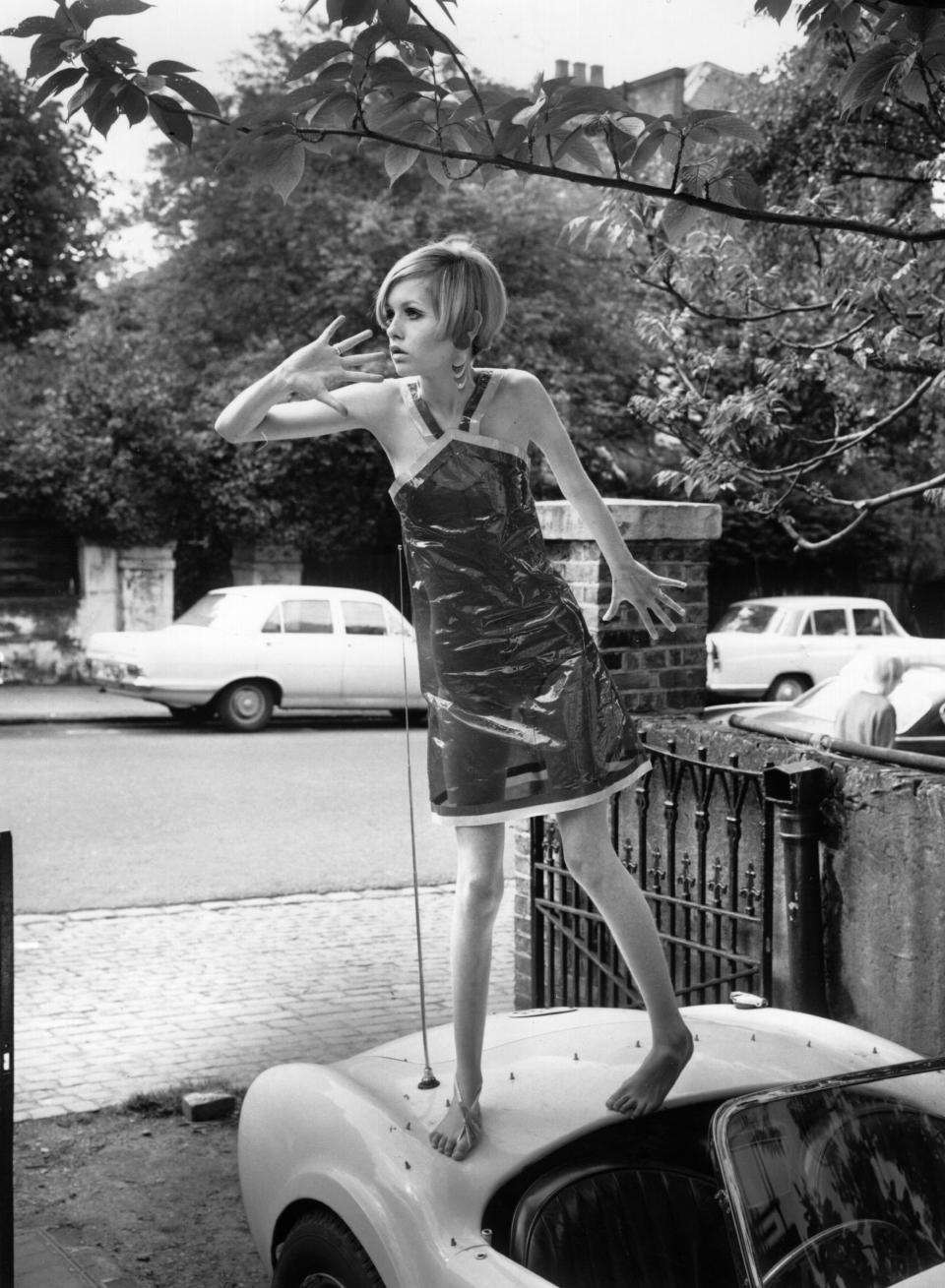 Twiggy models a transparent plastic halter neck dress while standing on a car.